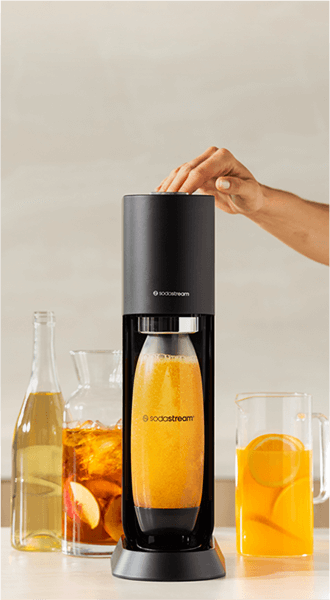 Czarny saturator z logo \'sodastream\' z butelką wypełnioną pomarańczowym napojem. Obok znajdują się dwa dzbanki z napojami i butelka z przezroczystym płynem.