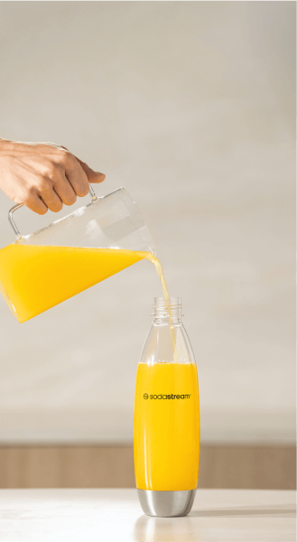 Osoba nalewa pomarańczowy napój z dzbanka do przezroczystej butelki z logo \'sodastream\'.