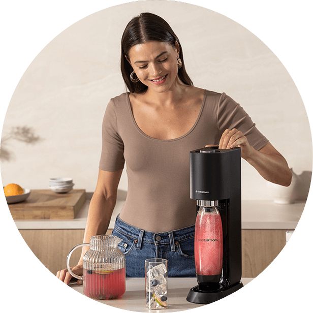 Osoba obsługuje czarny saturator marki SODASTREAM, w którym znajduje się różowy napój. Obok stoi dzbanek z napojem i szklanka z lodem i owocami.