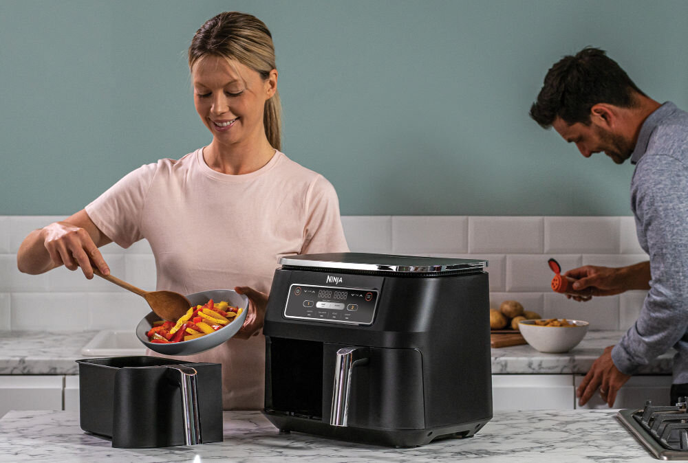 Air Fryer Frytkownica beztłuszczowa NINJA AF200EU z podwójnym koszem kobieta wsypująca do kosza warzywa mężczyzna przyprawiający potrawę frytkownica dwa kosze czarna obudowa tło kuchenne  pojemność 3,8 l łącznie daje 7,6 l dwa różne dania w mniejszych porcjach jedna większa porcja ceramiczna powłoka zapobieganie przywieraniu potraw kosze można myć w zmywarce szybkie wygodne sprzątanie funkcjonalność komfort w kuchni 