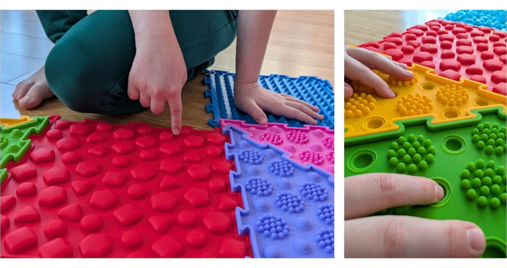 Mata sensoryczna ORTOPEDEK 910169 Zestaw mat (9 elementów) Wielokolorowy Dziecko dotyka dłońmi i palcami różnych tekstur mat sensorycznych, w tym czerwonej z figurami geometrycznymi, fioletowej z bąbelkami i zielonej z wypustkami.