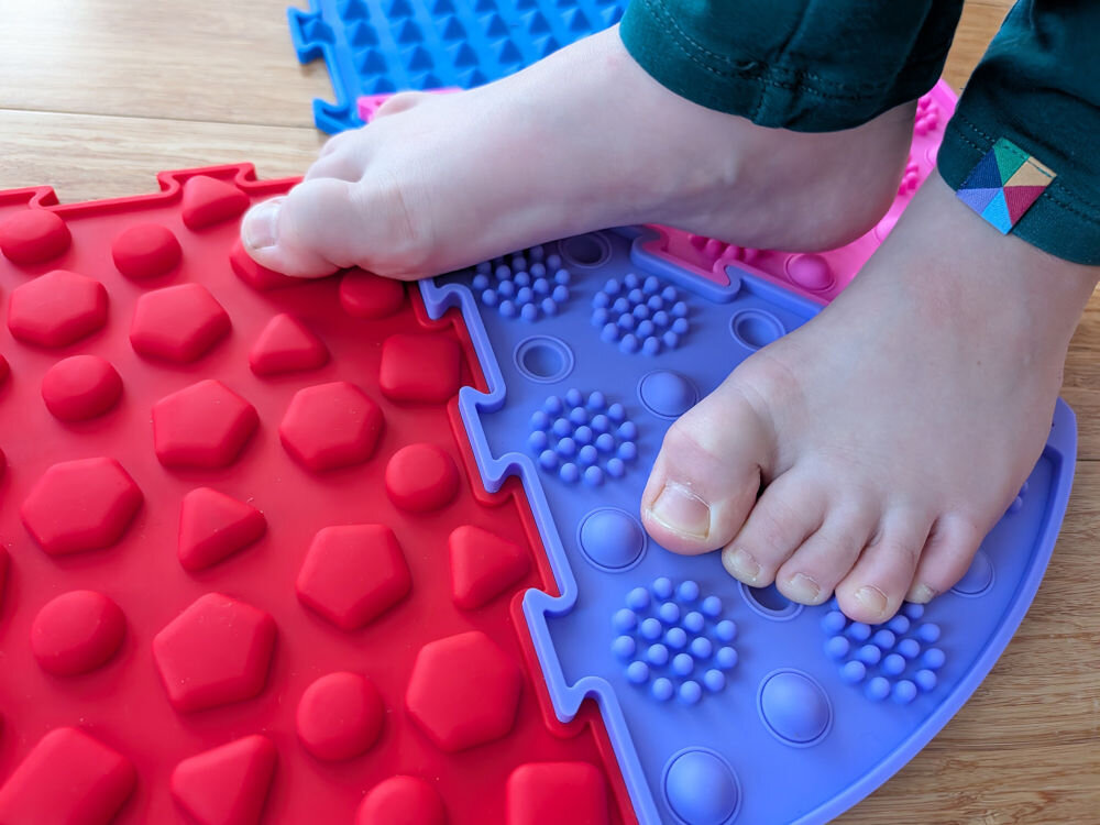 Mata sensoryczna ORTOPEDEK 910169 Zestaw mat (9 elementów) Wielokolorowy Bose stopy dziecka na kolorowej ścieżce zdrowia składającej się z czerwonej maty z wielokątami i fioletowej maty w kształcie choinki z masującymi bąbelkami.