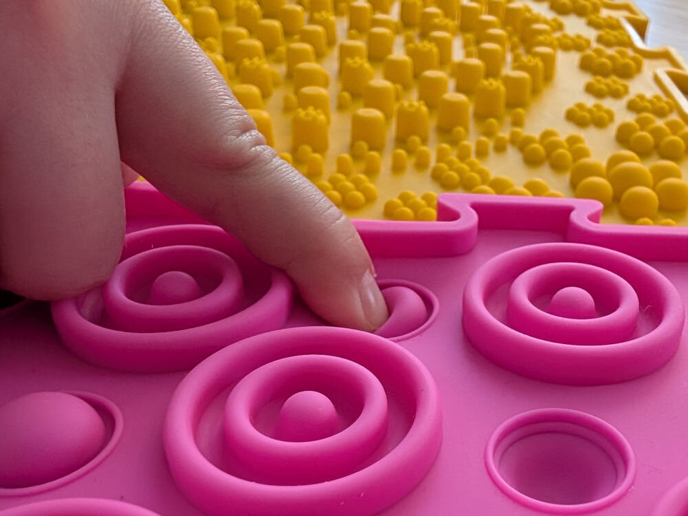 Mata sensoryczna ORTOPEDEK 910169 Zestaw mat (9 elementów) Wielokolorowy Zbliżenie na palec dziecka naciskający miękką, różową wypustkę na macie sensorycznej, w tle widoczna żółta mata z wysokimi elementami masującymi.