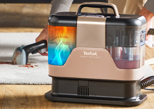 Odkurzacz piorący z widocznym logo \'Tefal Clean It Hot&Cold\' stoi na drewnianej podłodze obok dywanu, na którym ktoś używa końcówki do czyszczenia. Urządzenie ma przezroczyste zbiorniki z kolorowymi płynami.