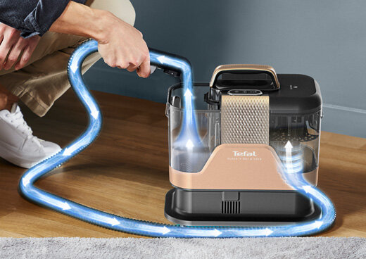 Osoba trzyma wąż od odkurzacza piorącego, który jest w kolorze czarno-złotym z widocznym logo \'Tefal\'. Wokół węża widoczne są niebieskie strzałki sugerujące przepływ powietrza.