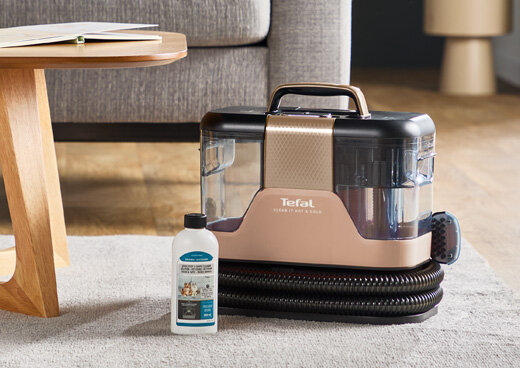 Na dywanie stoi odkurzacz piorący z widocznym logo \'Tefal Clean It Hot&Cold\', obok znajduje się butelka z płynem czyszczącym. W tle widać fragment stolika i kanapy.
