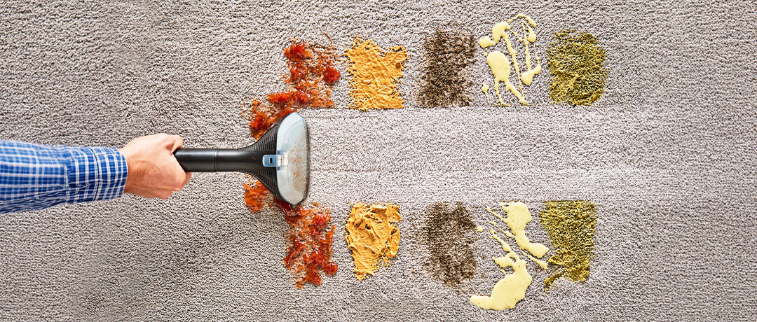 Ręka trzymająca odkurzacz piorący, który czyści dywan z różnorodnych plam i zabrudzeń, pozostawiając czysty pas pośrodku. Na dywanie widoczne są różne rodzaje zabrudzeń, takie jak sosy i okruchy.