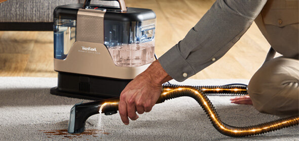 Osoba używa odkurzacza piorącego do czyszczenia plamy na dywanie. Na urządzeniu widoczne jest logo \'tefal\'.