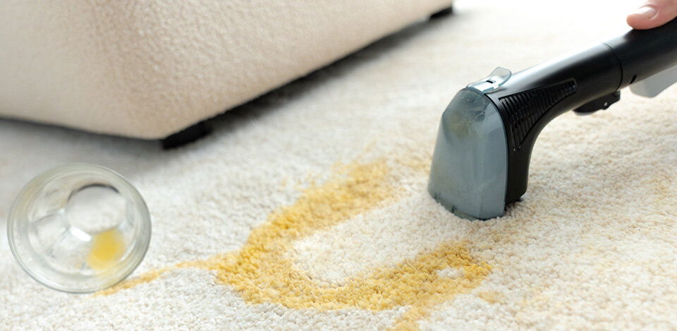 Na obrazie widać odkurzacz piorący czyszczący plamę na dywanie. Obok leży przewrócona szklanka z resztkami płynu.