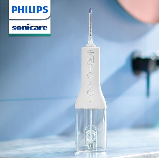 Na obrazie widoczny jest biały irygator z przezroczystym zbiornikiem na wodę, stojący na blacie. W lewym górnym rogu znajduje się logo \'PHILIPS sonicare\'.