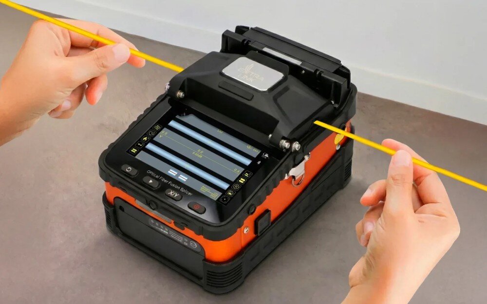 Spawarka światłowodowa EXTRALINK AI-5 Pro Pomarańczowo-czarna spawarka światłowodowa leżąca na blacie, do której dłonie wprowadzają z obu stron żółte włókna światłowodowe, na ekranie urządzenia widoczny jest interfejs z parametrami