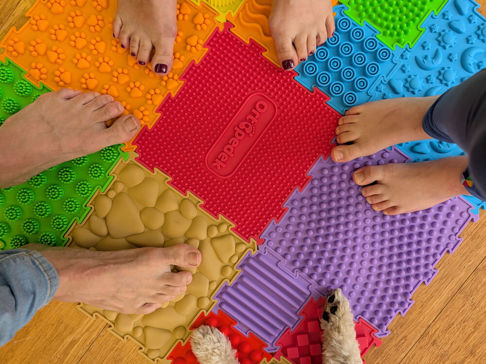 Mata sensoryczna ORTOPEDEK Zestaw mat 910176 (15 elementów) Wielokolorowy Widok z góry na grupę osób i psa stojących na dużym zestawie mat sensorycznych Ortopedek. Różnorodne faktury puzzli, w tym wzory kamieni, łap i wypustek w intensywnych kolorach.