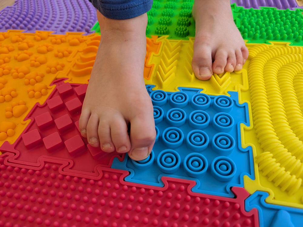 Mata sensoryczna ORTOPEDEK Zestaw mat 910176 (15 elementów) Wielokolorowy Bose stopy dziecka na kolorowej macie sensorycznej Ortopedek. Stopa naciska na niebieski element z wypustkami w kształcie kół, wspierając rozwój sensoryczny i prawidłową postawę.