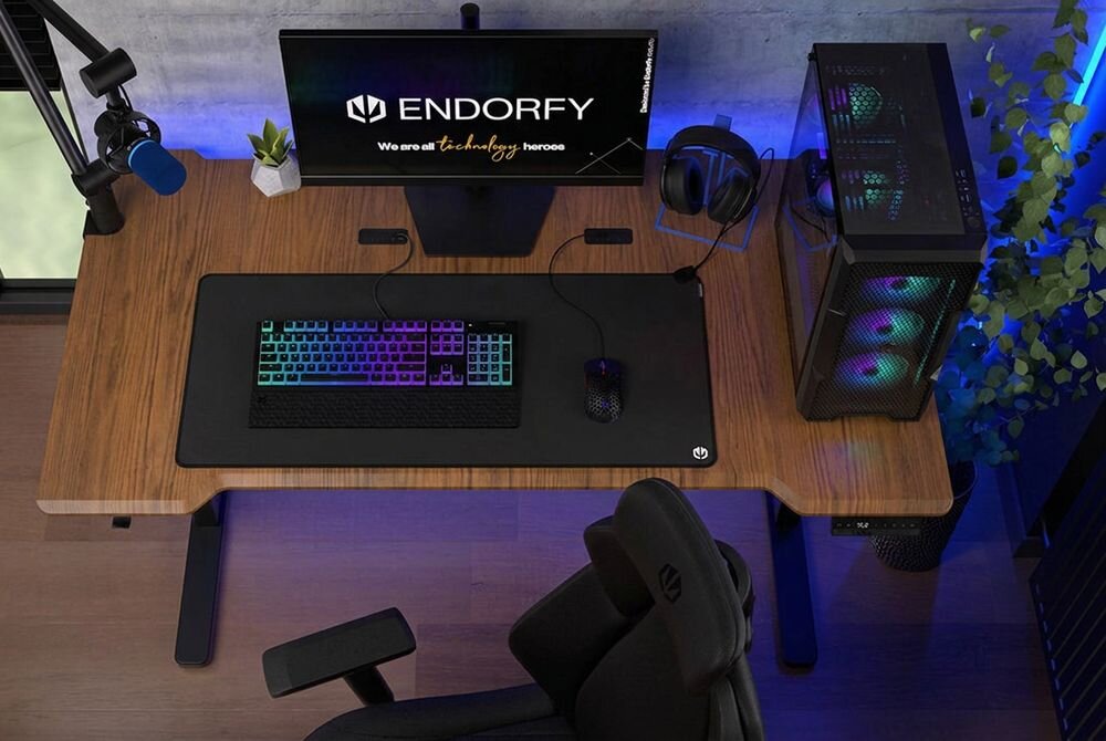 Na biurku znajduje się monitor z logo \'ENDORFY\' i tekstem \'We are all technology heroes\', klawiatura z podświetleniem RGB oraz mysz. Obok monitora stoi komputer z podświetleniem, a na biurku leży mikrofon i słuchawki.