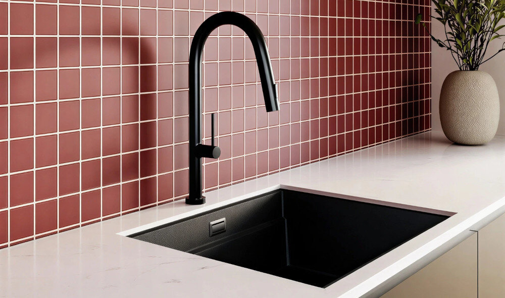 Czarna bateria kuchenna z wyciąganą wylewką zamontowana na białym blacie obok zlewu. W tle widoczna jest ściana z czerwonych płytek oraz dekoracyjna roślina w jasnej doniczce.