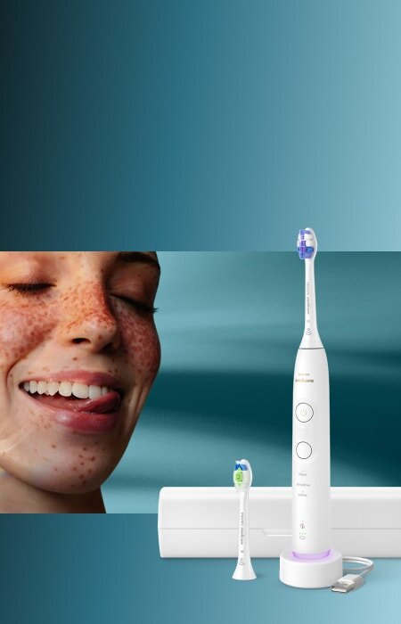 Zbliżenie na twarz uśmiechniętej kobiety, która językiem przesuwa po zębach. Obok szczoteczka soniczna z etui.