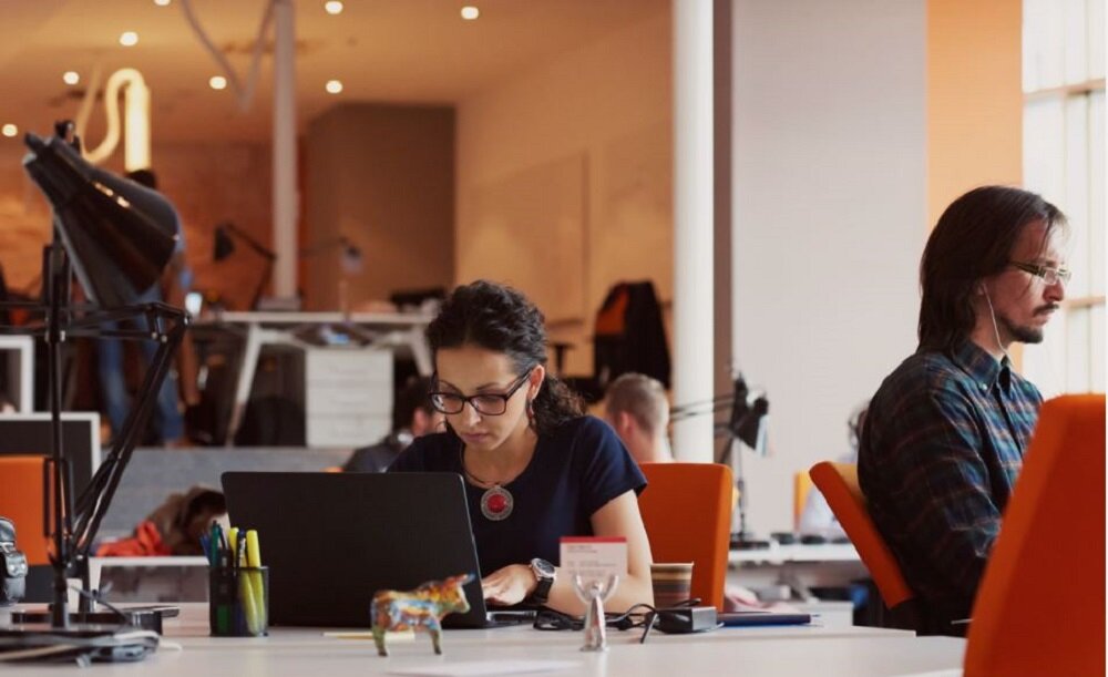 W biurze siedzą osoby przy biurkach, pracując na laptopach. W tle widać otwartą przestrzeń z biurkami i lampami.