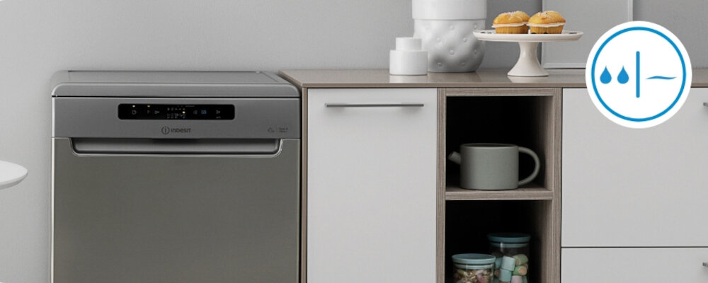 Zmywarka w kolorze srebrnym z panelem sterowania i logo \'Indesit\' obok szafki kuchennej. Na blacie obok znajdują się filiżanki i patera z babeczkami, a na ścianie widoczna jest ikona z kroplami wody.