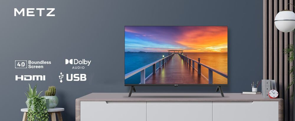 Telewizor METZ stoi na szafce, wyświetlając obraz molo prowadzącego w stronę zachodzącego słońca. Obok telewizora znajdują się logotypy: METZ, 4.0 Boundless Screen, Dolby Audio, HDMI, USB.