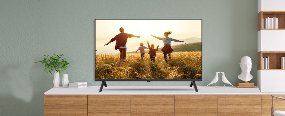 Telewizor wyświetla obraz przedstawiający cztery osoby biegnące przez pole o zachodzie słońca. Urządzenie stoi na białej szafce, obok której znajduje się roślina w doniczce i kilka książek.