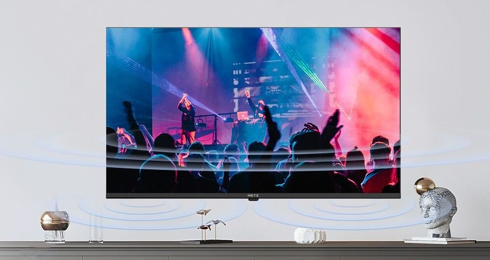 Telewizor METZ wyświetla kolorową scenę koncertową z widocznymi sylwetkami ludzi. Na półce pod telewizorem znajdują się dekoracyjne przedmioty, w tym rzeźba głowy i mała roślina.