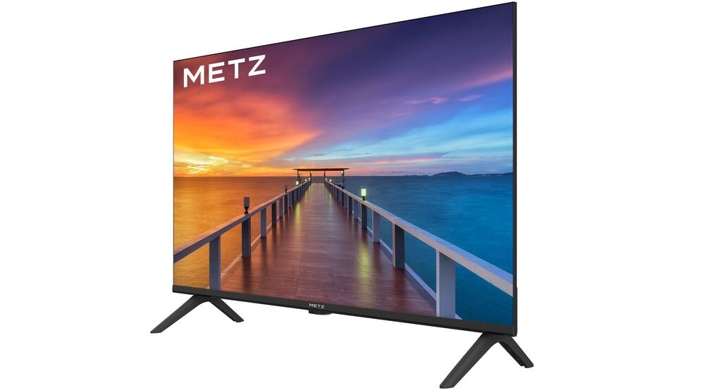Telewizor z logo METZ na ekranie, wyświetlający obraz molo prowadzącego w stronę zachodzącego słońca nad wodą. Urządzenie stoi na dwóch nóżkach.
