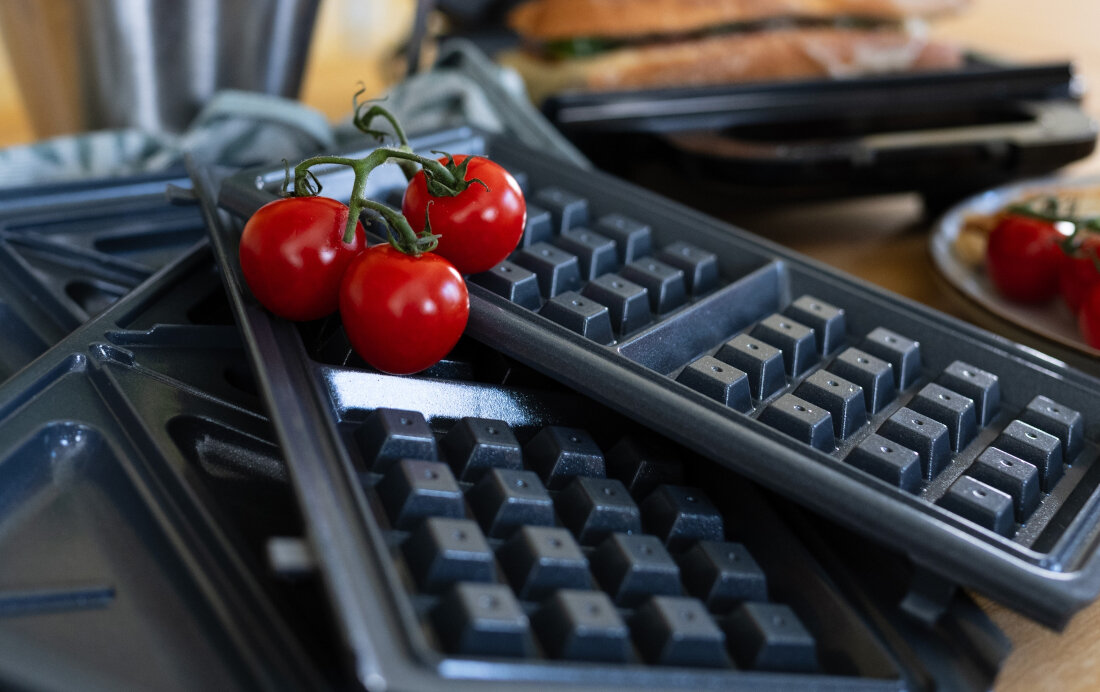 Na obrazie widoczne są metalowe płyty do opiekacza z wzorem do gofrów, na których leżą gałązki pomidorów koktajlowych. W tle znajduje się kanapka i inne elementy kuchenne.