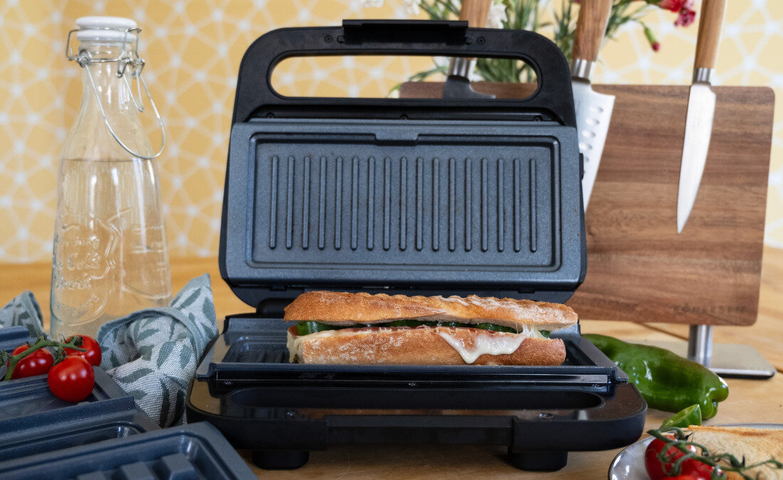 Na blacie kuchennym znajduje się otwarty opiekacz z kanapką w środku. W tle widać deskę do krojenia z nożami oraz butelkę z wodą i pomidory koktajlowe.