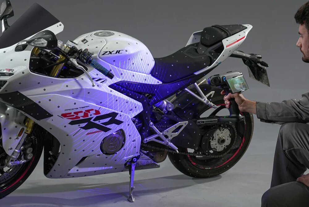 Skaner 3D CREALITY Sermoon S1 Osoba w szarym kombinezonie, kucając, skanuje prawy bok białego motocykla sportowego ręcznym skanerem 3D, który wyświetla siatkę niebieskich linii na boku pojazdu, prędkość, klatki na sekundę