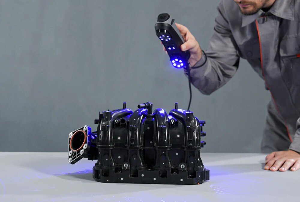 Skaner 3D CREALITY Sermoon S1 Mężczyzna w szarym kombinezonie roboczym trzyma w prawej ręce skaner 3D, z którego wydobywa się intensywne niebieskie światło, oświetlając czarny, plastikowy kolektor dolotowy silnika leżący na stole, przygotowanie modelu, praktyczność