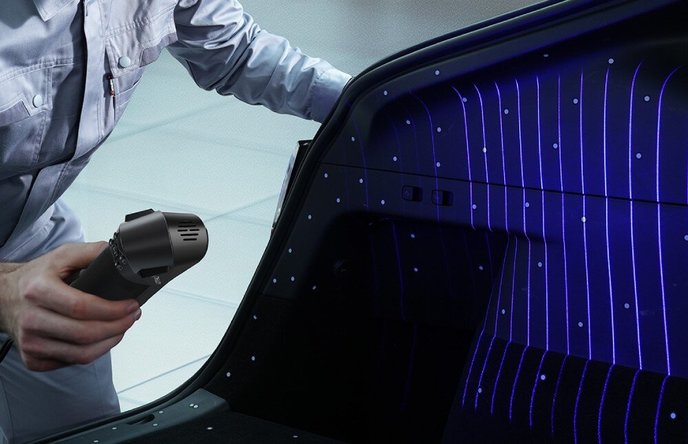 Skaner 3D CREALITY CR-Scan Raptor Pro Osoba w szarym ubraniu roboczym trzyma urządzenie, które rzuca siatkę niebieskich linii laserowych oraz jasne punkty na czarne wnętrze bagażnika samochodu, tryby, lasery, obsługa wielkości