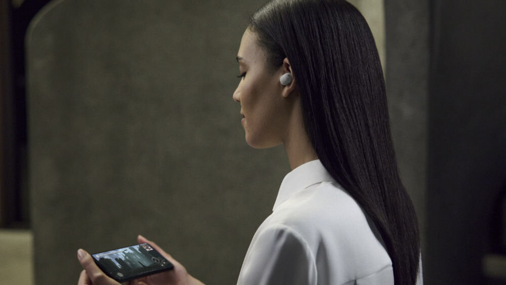 Słuchawki dokanałowe SONY WF-1000XM6 ANC Kobieta w białej koszuli oglądająca wideo na smartfonie, korzystająca ze srebrnych słuchawek dousznych Sony WF-1000XM6.