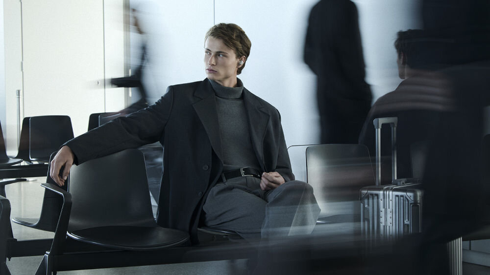 Słuchawki dokanałowe SONY WF-1000XM6 ANC Mężczyzna siedzący w poczekalni lotniska ze srebrną słuchawką Sony WF-1000XM6 w uchu; obok widoczna walizka.