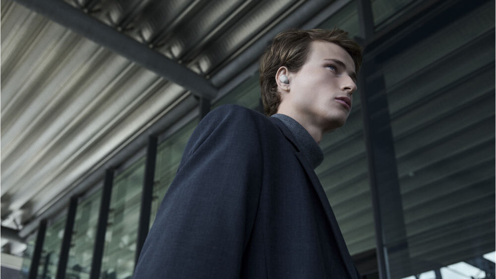 Słuchawki dokanałowe SONY WF-1000XM6 ANC Mężczyzna w ciemnej marynarce na tle nowoczesnej architektury, prezentujący srebrną słuchawkę Sony WF-1000XM6.