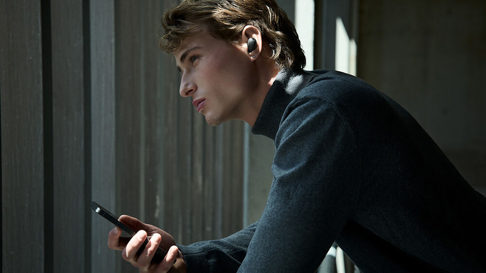 Słuchawki dokanałowe SONY WF-1000XM6 ANC Mężczyzna w czarnym golfie patrzący przez okno i trzymający smartfona, z czarną słuchawką Sony WF-1000XM6 w uchu.