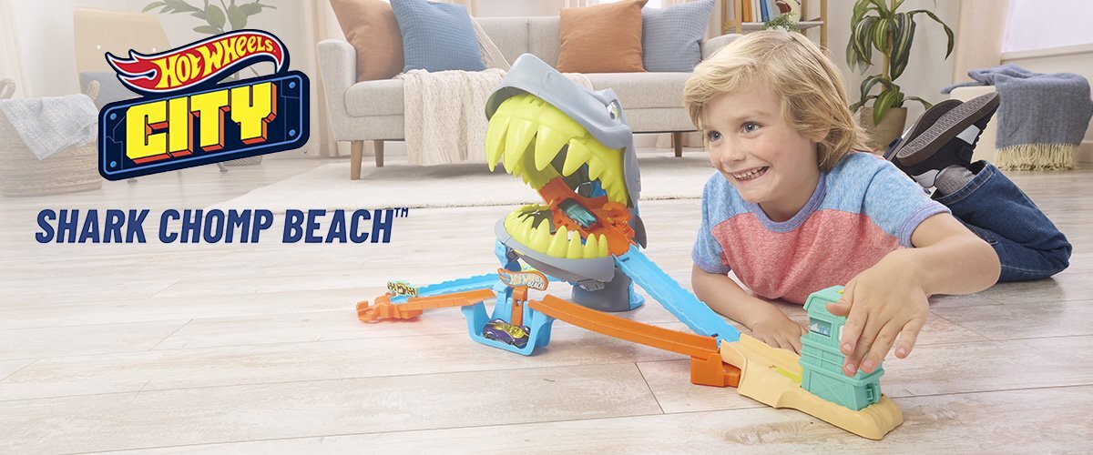 Dziecko bawi się torem Hot Wheels z dużą figurką rekina. Obok widoczne jest logo Hot Wheels City oraz napis \'Shark Chomp Beach\'.