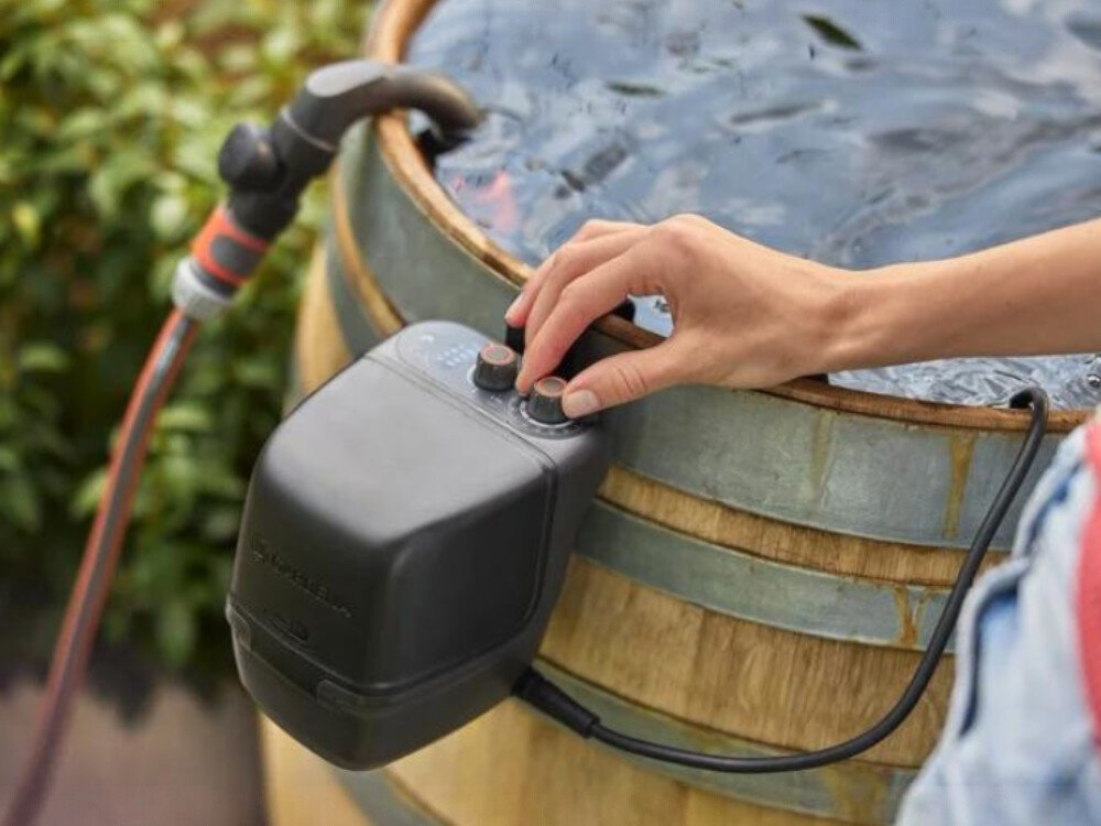 Osoba obsługuje czarną pompę do wody zamontowaną na drewnianej beczce z wodą. W tle widoczny jest wąż ogrodowy podłączony do pompy.
