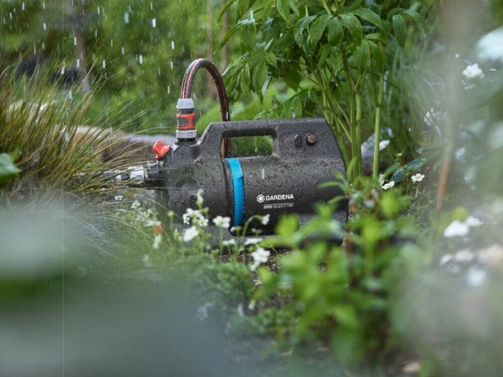 Na obrazie widoczna jest pompa do wody marki Gardena, umieszczona w ogrodzie wśród roślin. Na urządzeniu widnieje napis \'GARDENA 3000/3 18V\'.