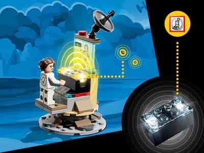 Zestaw LEGO przedstawia postać siedzącą przy konsoli z ekranem i anteną satelitarną, na tle niebieskiego krajobrazu. W prawym górnym rogu widoczna jest ikona z symbolem baterii, a obok niej element LEGO z podświetleniem.