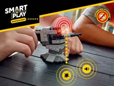 Na stole znajduje się model statku kosmicznego z klocków, wokół którego widoczne są efekty graficzne symbolizujące dźwięk i światło. W lewym górnym rogu widnieje logo \'SMART PLAY\', a w prawym górnym rogu okrągły znak z przekreślonym telefonem i napisem \'ZAKAZ BEZ EKRANU\'.