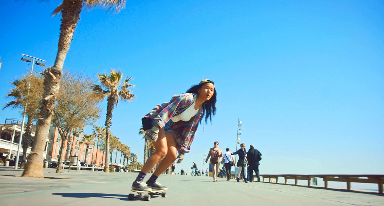 Dziewczyna na deskorolce jeździ środkiem ulicy, mając molo po lewej stronie i palmy po prawej.