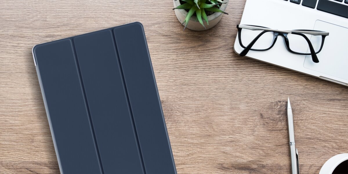 Na drewnianym biurku leży granatowe etui na tablet, obok którego znajdują się okulary, długopis, filiżanka kawy i laptop. W tle widoczna jest mała roślina w doniczce.