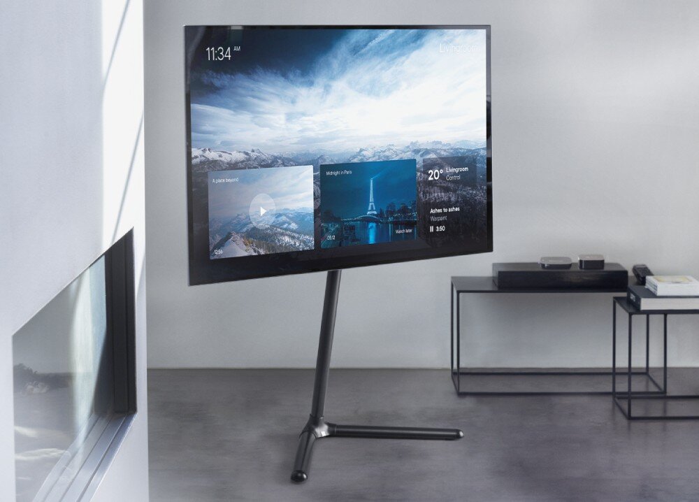 Stojak podłogowy ONE FOR ALL Zdjęcie przedstawia duży telewizor zamontowany na czarnym stojaku podłogowym z jedną nogą wygiętą pod kątem i rozwidloną podstawą. Telewizor znajduje się w jasnym pokoju o betonowej podłodze, a na ekranie widoczne są kafelki interfejsu z pejzażami i zegarem. W tle po prawej stronie stoją dwa niskie, metalowe stoliki kawowe, na których leżą drobne przedmioty elektroniczne