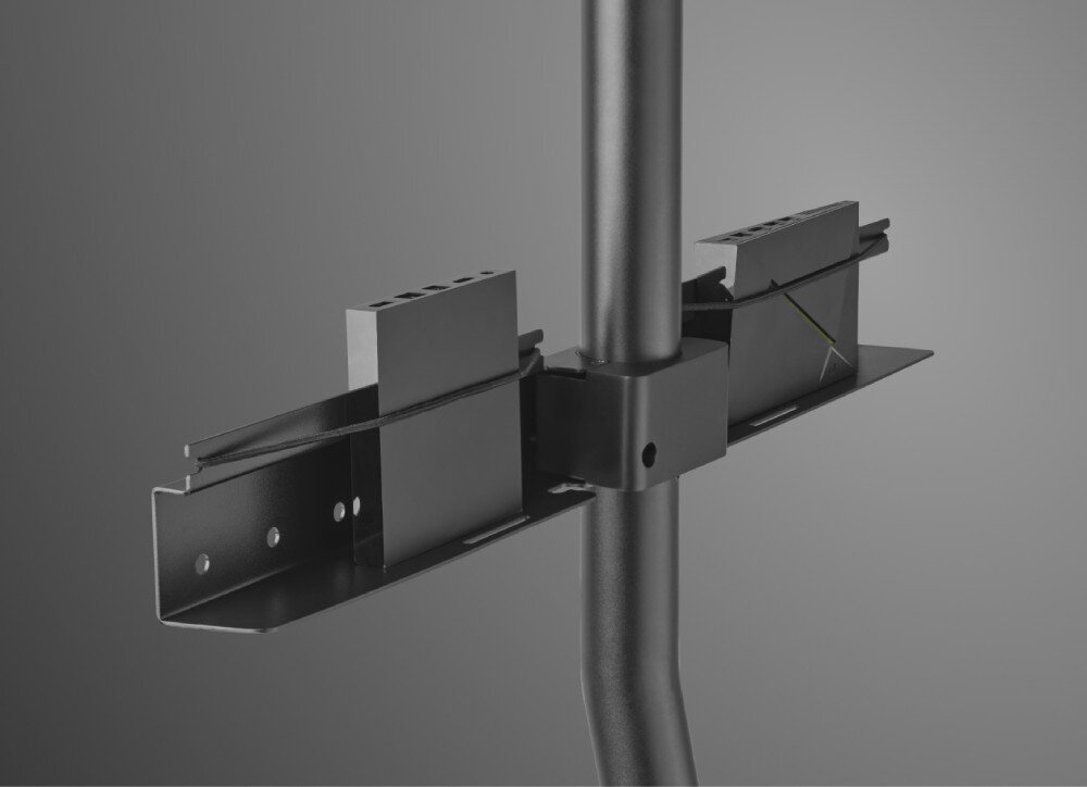 Stojak podłogowy ONE FOR ALL Zbliżenie na środkową część czarnego stojaka, gdzie pod ramą montażową telewizora znajduje się metalowa półka. Na półce ustawione są dwa małe, prostokątne urządzenia elektroniczne przypominające dekodery, które są przytrzymywane przez cienką, czarną linkę zabezpieczającą