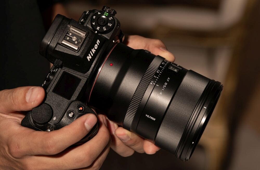 Osoba trzyma aparat fotograficzny z obiektywem marki VILTROX AF 85mm f/1.4, zamontowanym na korpusie Nikon. Na obiektywie widoczne są napisy \'VILTROX\' i \'AF 85/1.4\'.