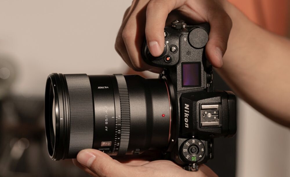 Osoba trzyma aparat fotograficzny z zamontowanym obiektywem, na którym widoczny jest napis \'VILTROX AF 85mm f/1.4 Pro\'. Na korpusie aparatu widnieje logo \'Nikon\'.