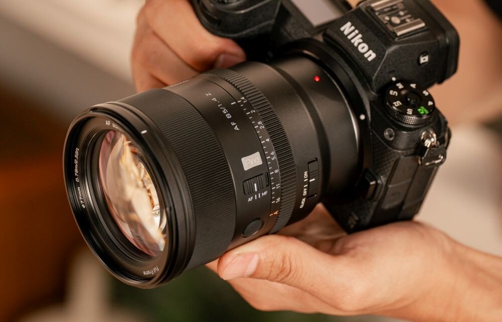 Osoba trzyma aparat fotograficzny z zamontowanym obiektywem VILTROX AF 85mm f/1.4 Pro. Na aparacie widoczne jest logo \'Nikon\'.