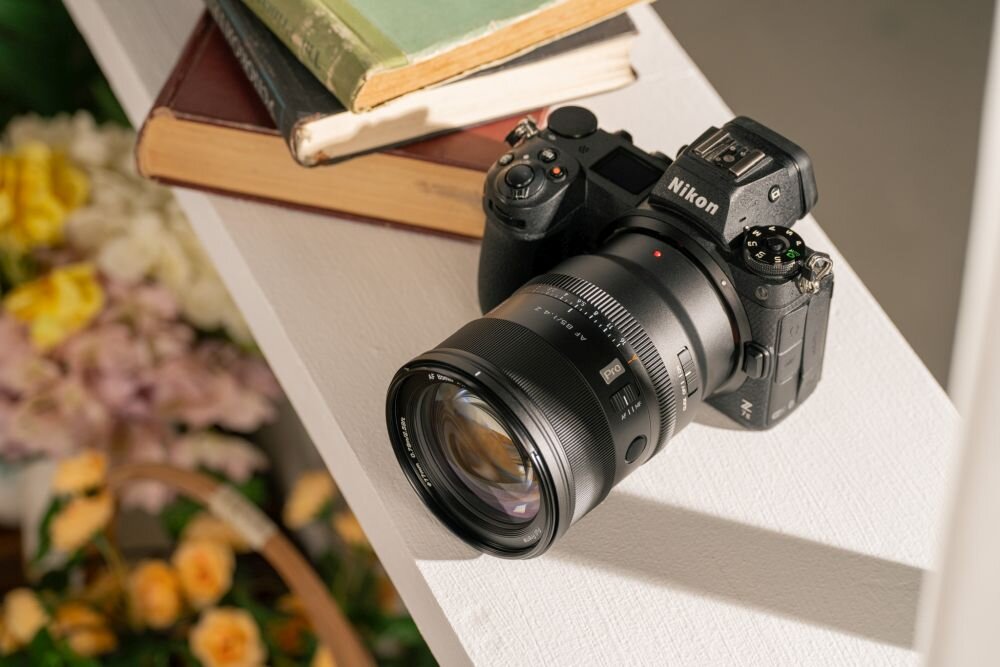Aparat fotograficzny z obiektywem leży na białej półce obok stosu książek. Na aparacie widoczne jest logo \'Nikon\'.