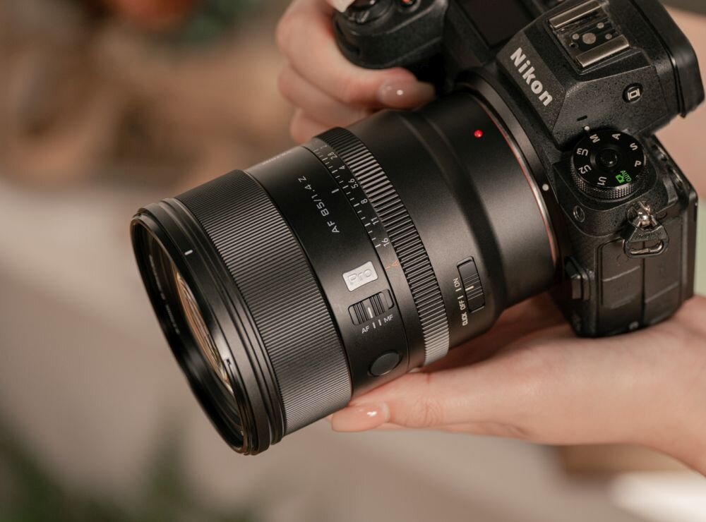 Osoba trzyma aparat fotograficzny z zamontowanym obiektywem VILTROX AF 85mm f/1.4 Pro Nikon Z. Na aparacie widoczne jest logo \'Nikon\'.