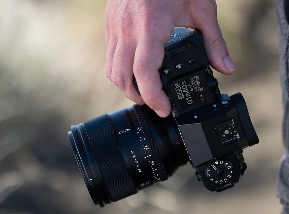 Osoba trzyma aparat fotograficzny z zamontowanym obiektywem VILTROX AF 56mm f/1.2. Na wyświetlaczu aparatu widoczne są różne ustawienia i parametry.